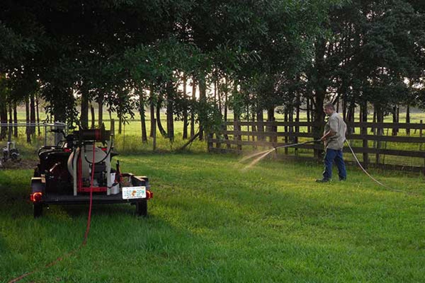 Man spraying lawn with a sprayer attached to a trailer in front of a fence and trees.