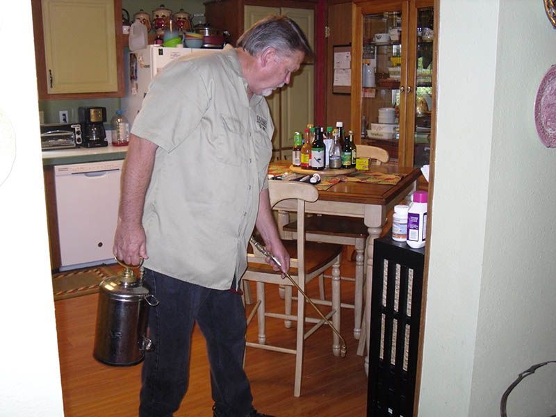 Man in beige shirt and jeans points with a stick in a kitchen.