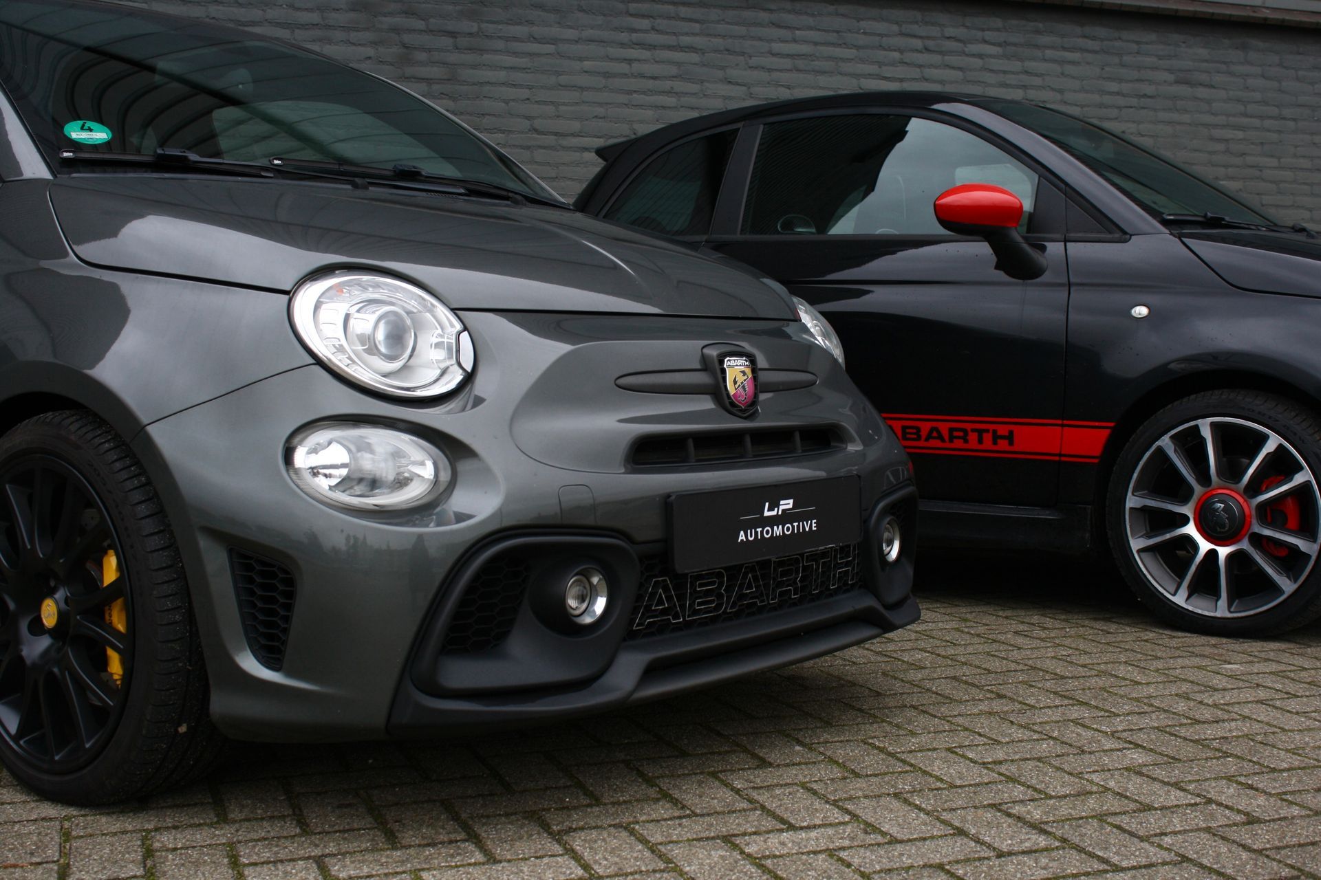 Twee donkergrijze en zwarte Abarth 500-auto's geparkeerd op een bakstenen trottoir.