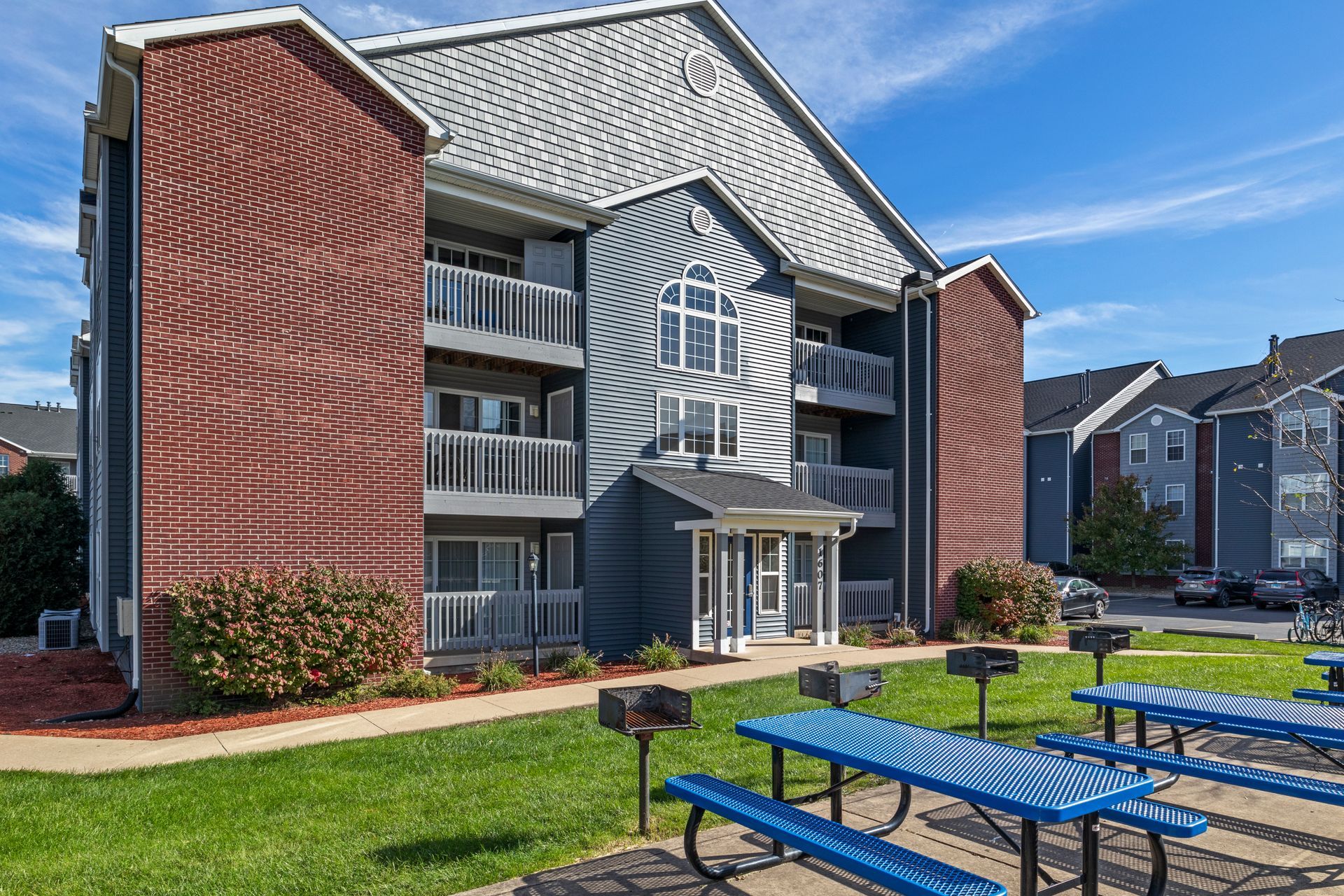 A large apartment building with blue picnic tables and grills in front of it.