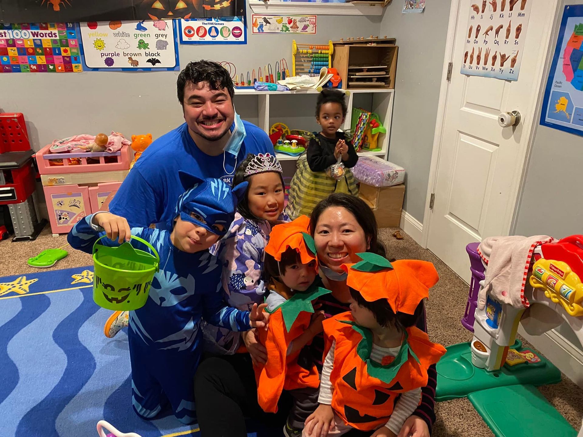 Group of children and adults in a playroom wearing Halloween costumes, smiling.