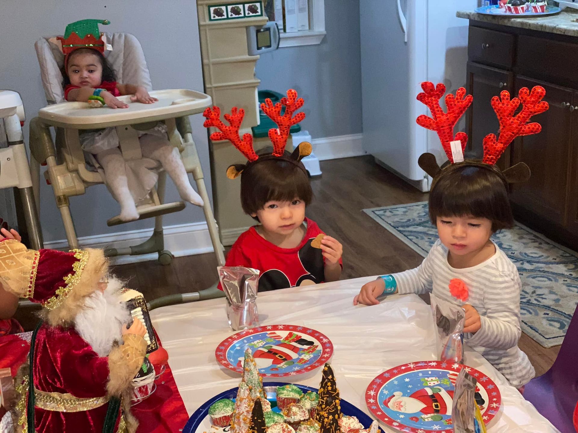 Children at a Christmas table wearing festive headbands and eating. One child in a high chair. Santa decoration.