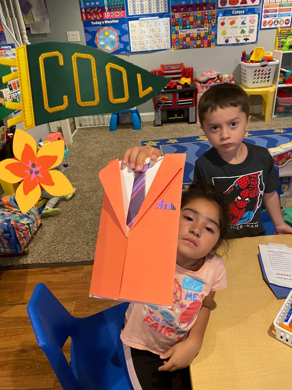 Two children holding a paper suit craft in a classroom. The boy looks on.
