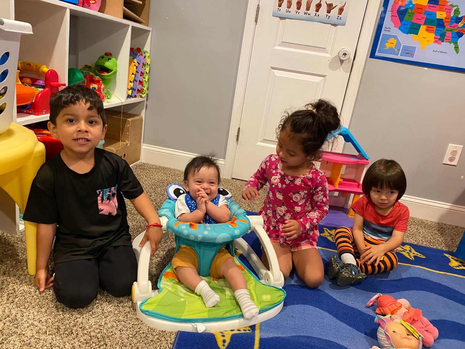 Four children in a play area; one in a walker, smiling. Other children looking at the camera.