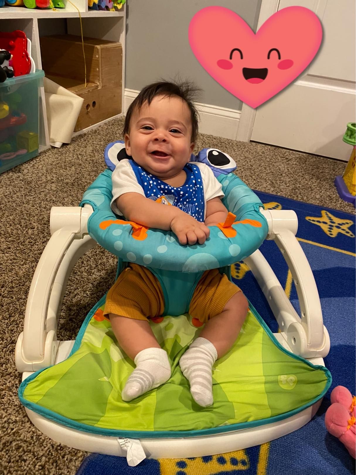 Smiling baby in a blue and green activity seat, wearing a bib, sitting on a carpeted floor. A pink heart is overlaid.