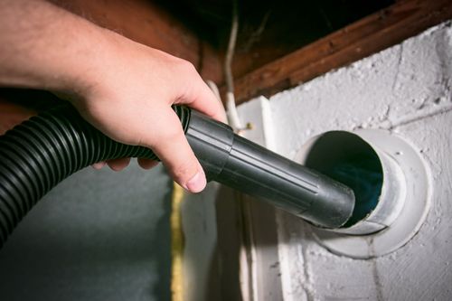 A person uses a vacuum hose attachment to clean a circular dryer vent opening on a white wall.