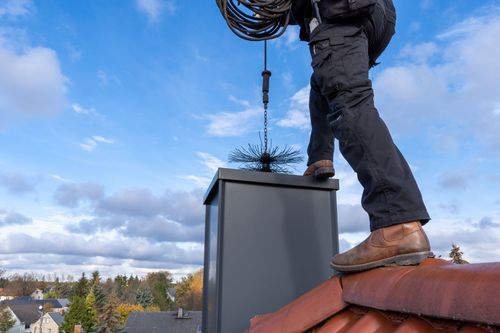 A person in dark work clothes cleans a rooftop chimney with a rotating brush tool under a partly cloudy blue sky.