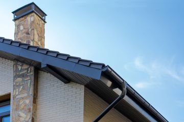 A stone chimney and dark tiled roof with a matching gutter system against a clear blue sky.