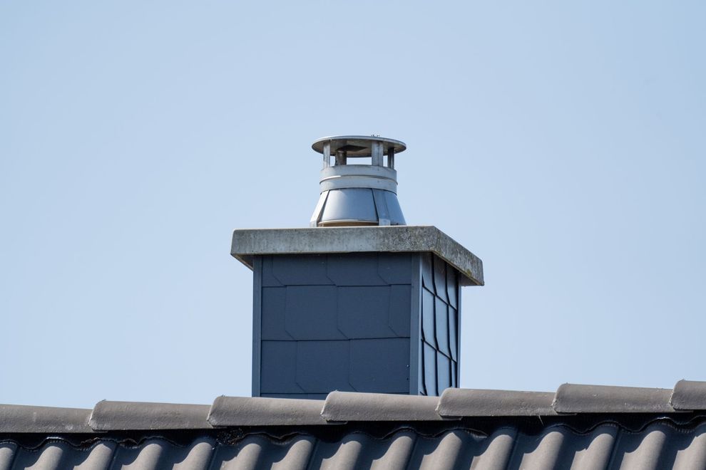 A grey tiled chimney with a metal cap, rising from a grey tiled roof against a clear blue sky.
