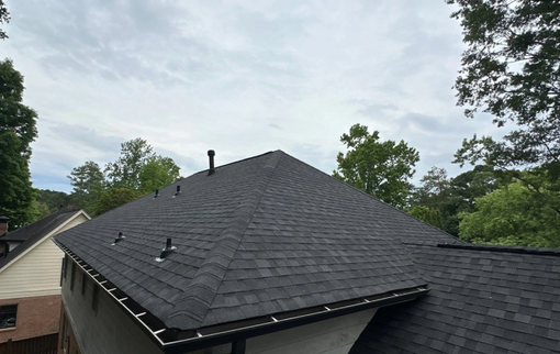 Dark gray shingle roof on a house, under a cloudy sky. Trees are visible in the background.
