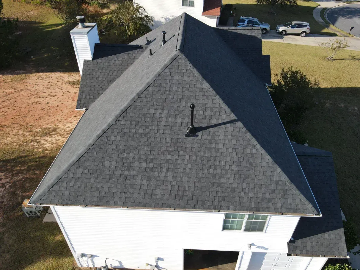 A house with a dark gray shingled roof, seen from above, with chimney and vent pipes.