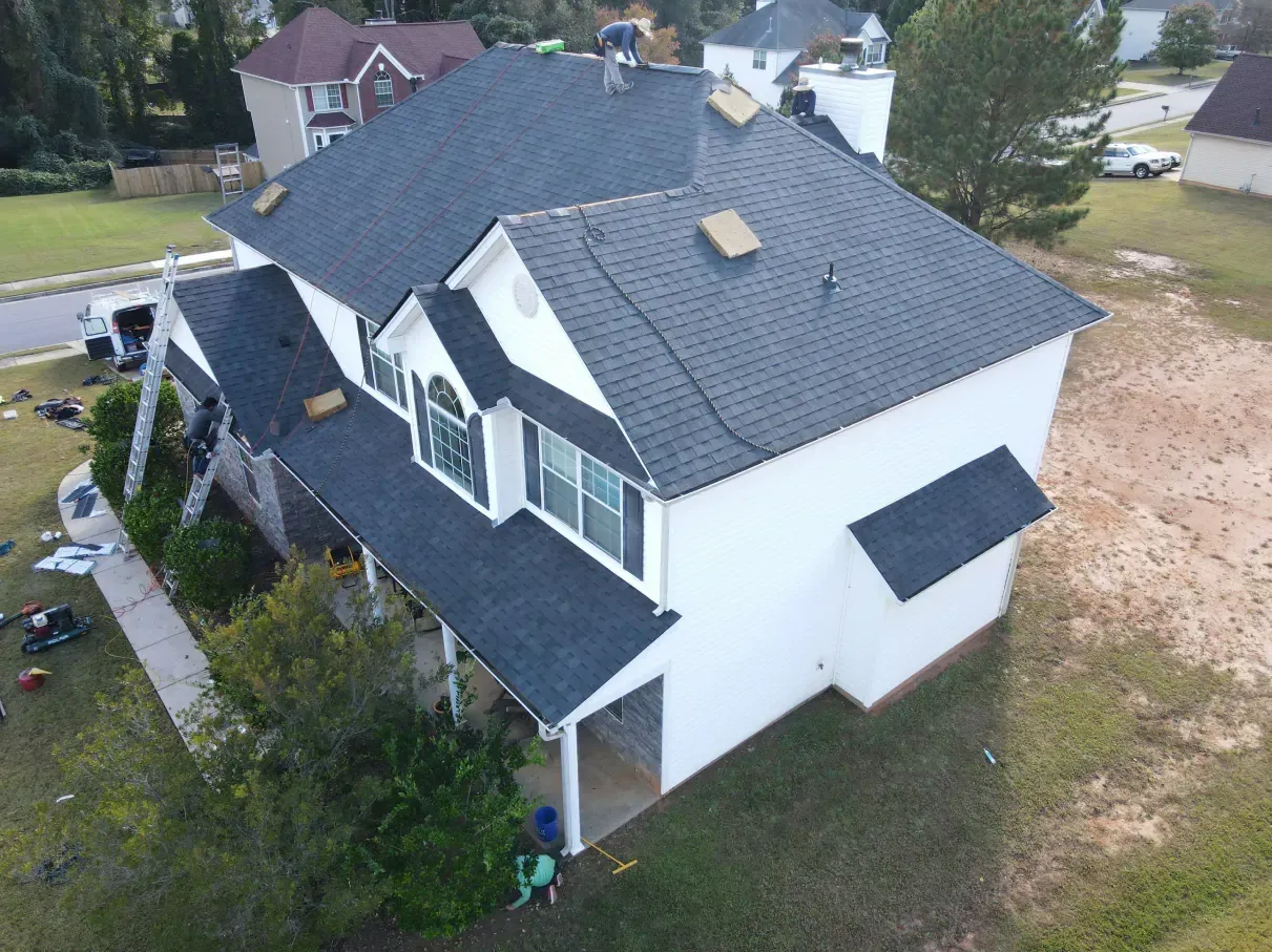Two-story house with dark gray roof being worked on by roofing crew. White siding, green grass, and trees.