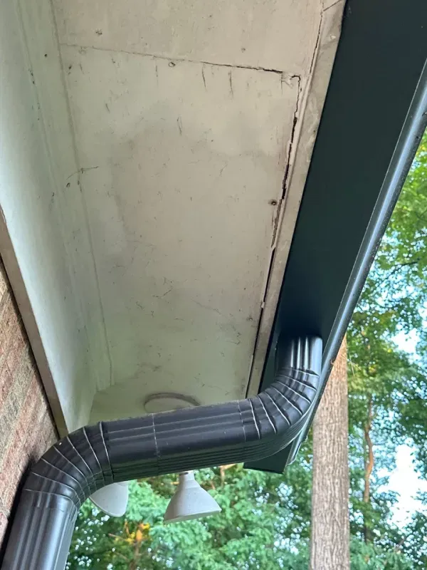 Dark guttering and downspout on a house, with white ceiling and brick wall visible.