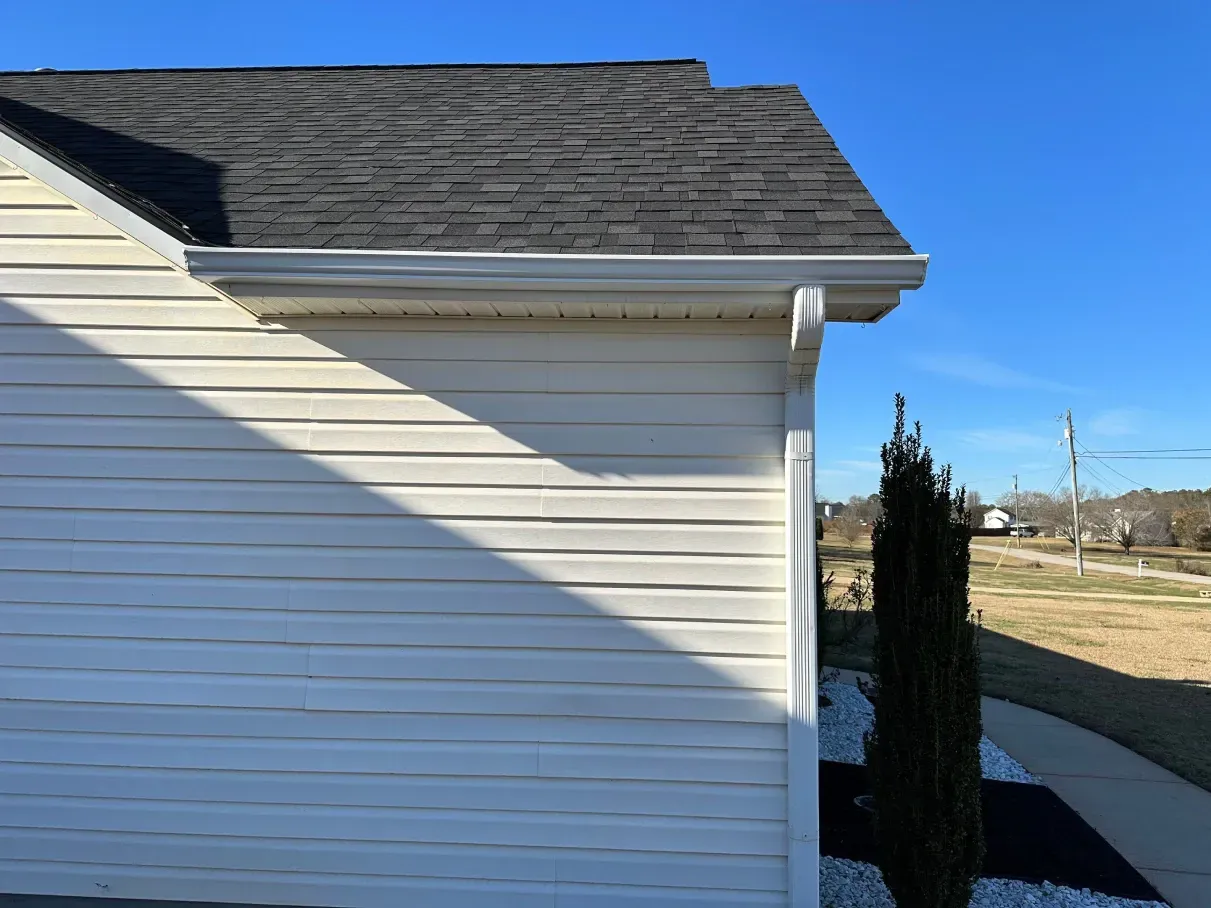 Side view of a house with white siding, dark roof, and white gutters against a blue sky.