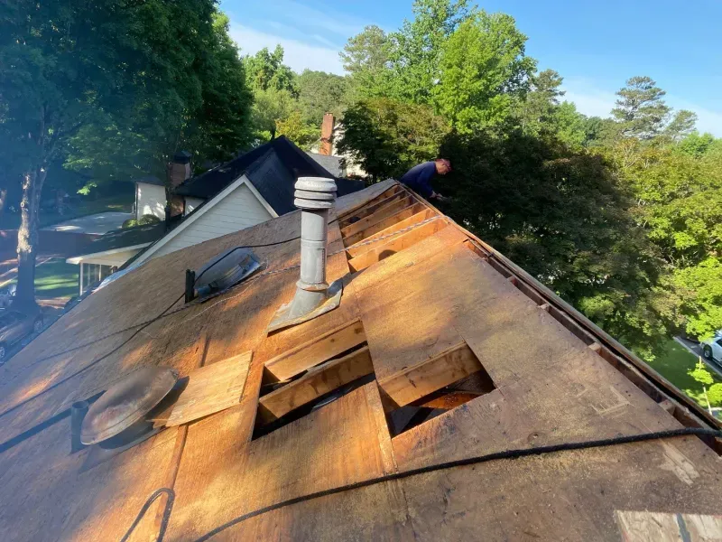 Roofer working on a roof, daylight. Damaged wooden boards, chimney, and blue sky in the background.