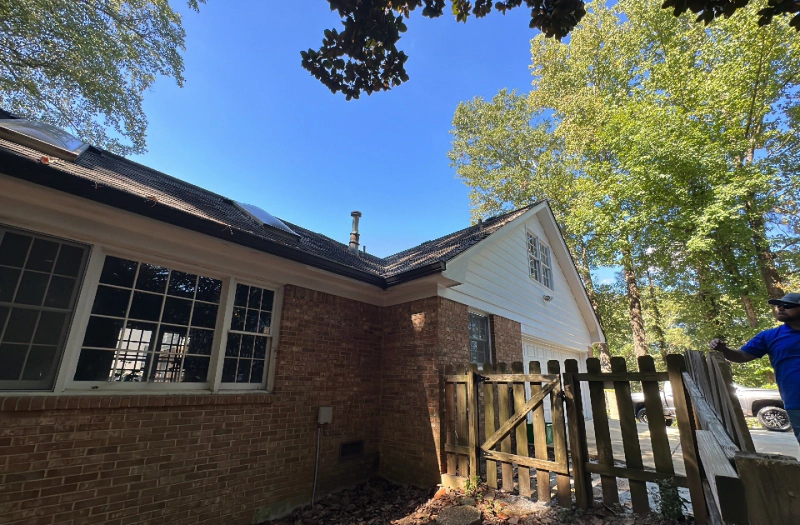 Side view of a brick house with a dark roof, wooden fence, and blue sky. A person stands nearby.