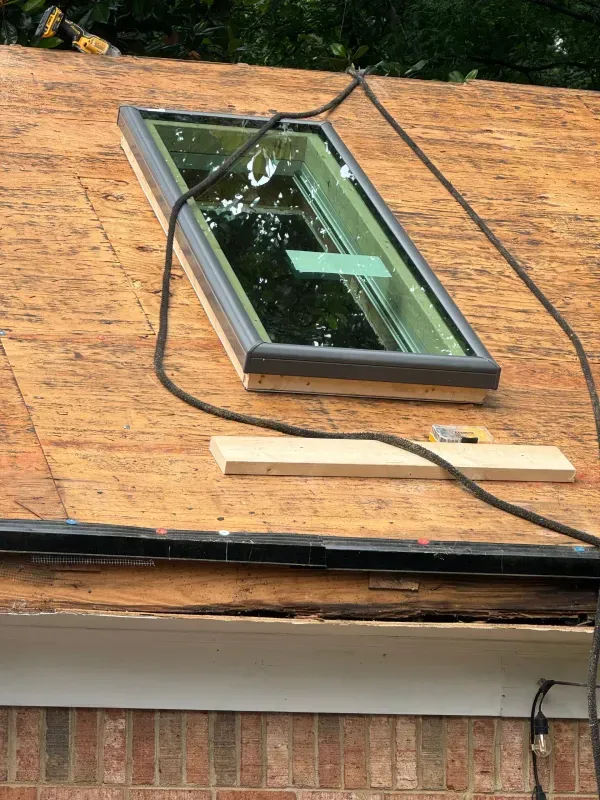 Skylight installed on a roof, black frame, gray sealant, brown wood, black cable, white building.