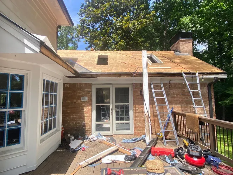 Roof construction in progress on a brick home with skylights, on a wooden deck, with ladders.