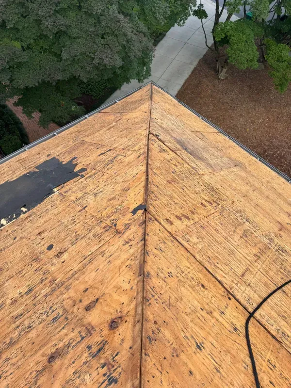 Overhead view of a roof with exposed wooden sheathing, some black underlayment, and a driveway in the background.