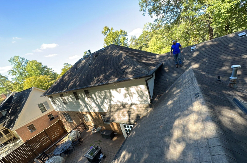 A person on a roof with dark shingles on a sunny day. A building with a deck and trees is visible.