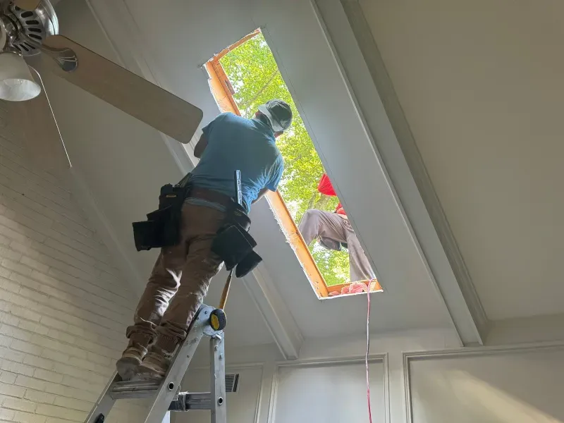 Person on ladder installing a skylight in a room with white walls and a ceiling fan.