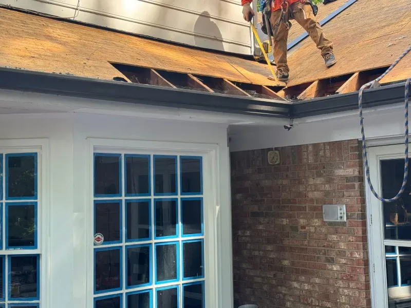 Roofer working on a roof section with a safety harness. Windows below with brick exterior.