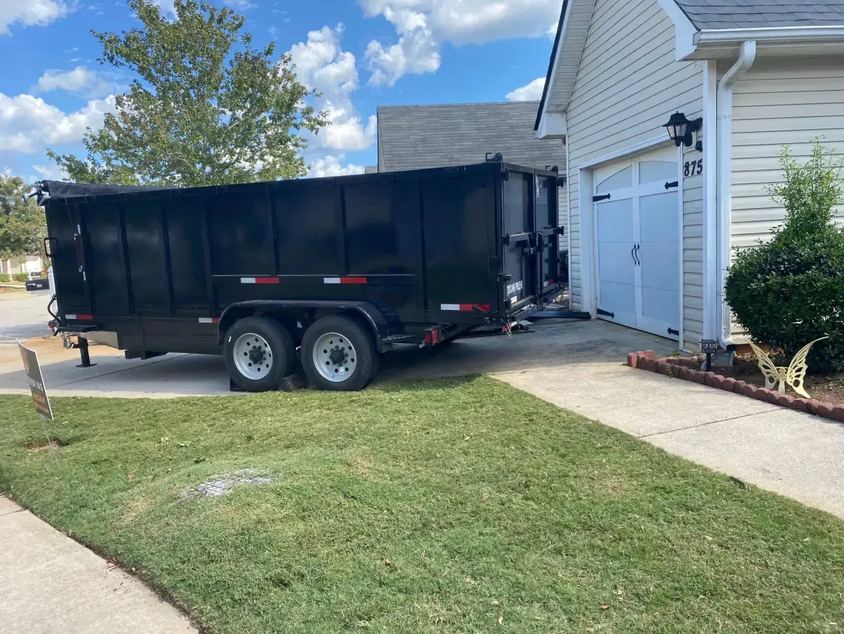Black dumpster trailer parked on a driveway beside a garage, green grass and street visible.
