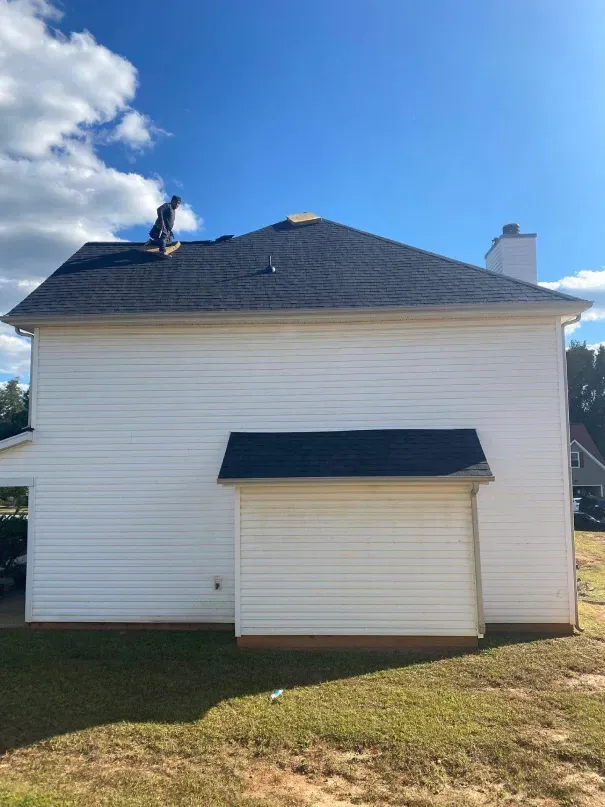 Person on a roof installing shingles on a white house with a blue sky background.