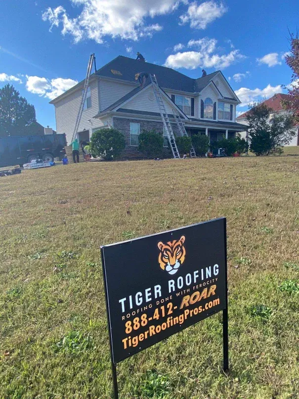 Tiger Roofing sign in front of a house with ladders on the roof, under a blue sky.