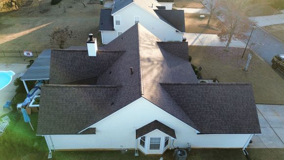 Person on roof replacing shingles on a white house with blue sky and clouds.