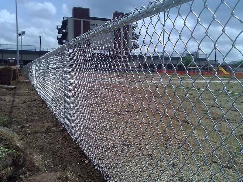 A chain link fence surrounds a field; a multi-story brick building is in the background.
