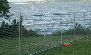 Chain-link fence along a grassy shoreline with a body of water and distant buildings.