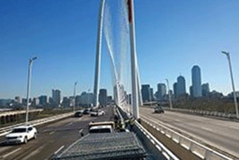 Cars on a bridge with a large white arch, tall buildings in background on a clear day.