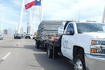 White truck carrying metal sheets on a bridge with the Texas flag waving in the background.