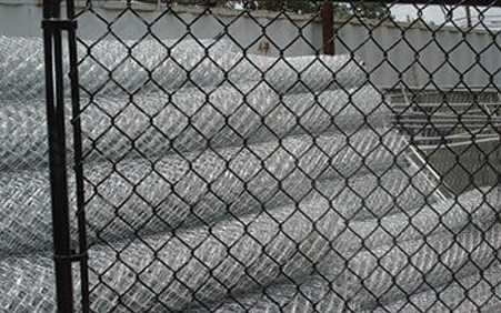 Black chain-link fence in front of stacked rolls of chain-link fencing, possibly at a construction site.