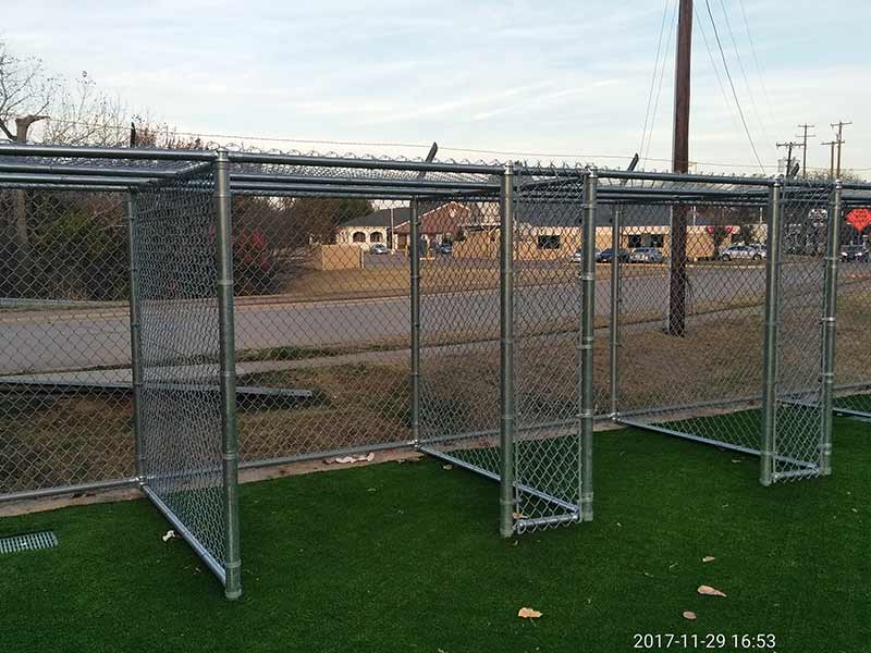 Chain-link dugout shelters on a green turf field. A road and buildings are visible in the background.