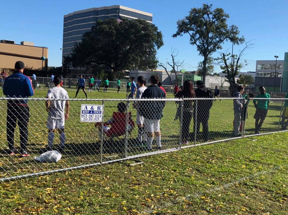 Soccer game in progress, viewed from behind a chain-link fence. Spectators watch on a sunny day.