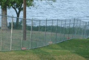 Chain-link fence on green grass next to a body of water, tree in the background.