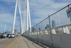 A white suspension bridge in Dallas, TX, with chain-link fence, vehicles, and city skyline in background.