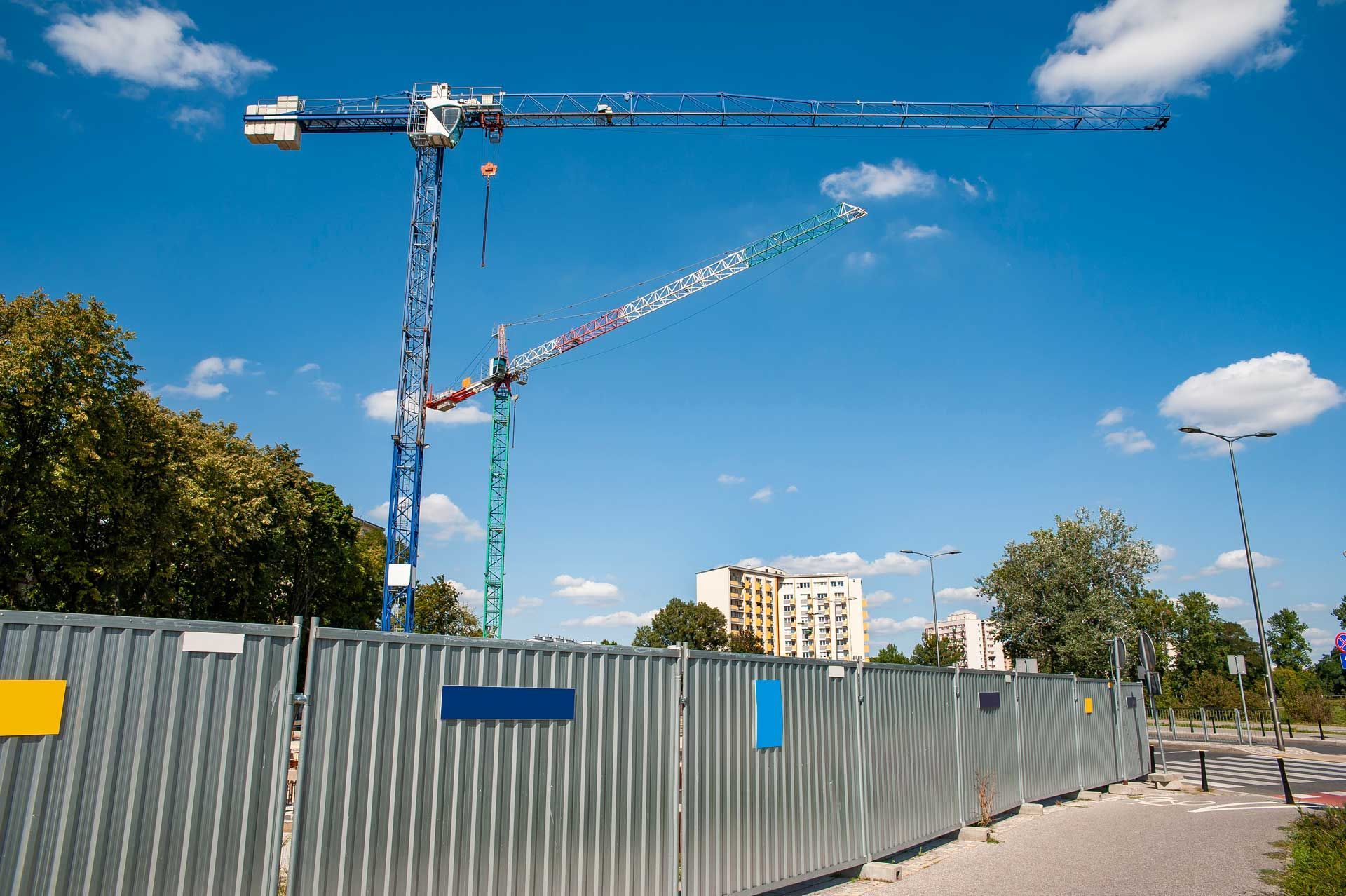 Two tall cranes rise over a fenced construction site on clear day.