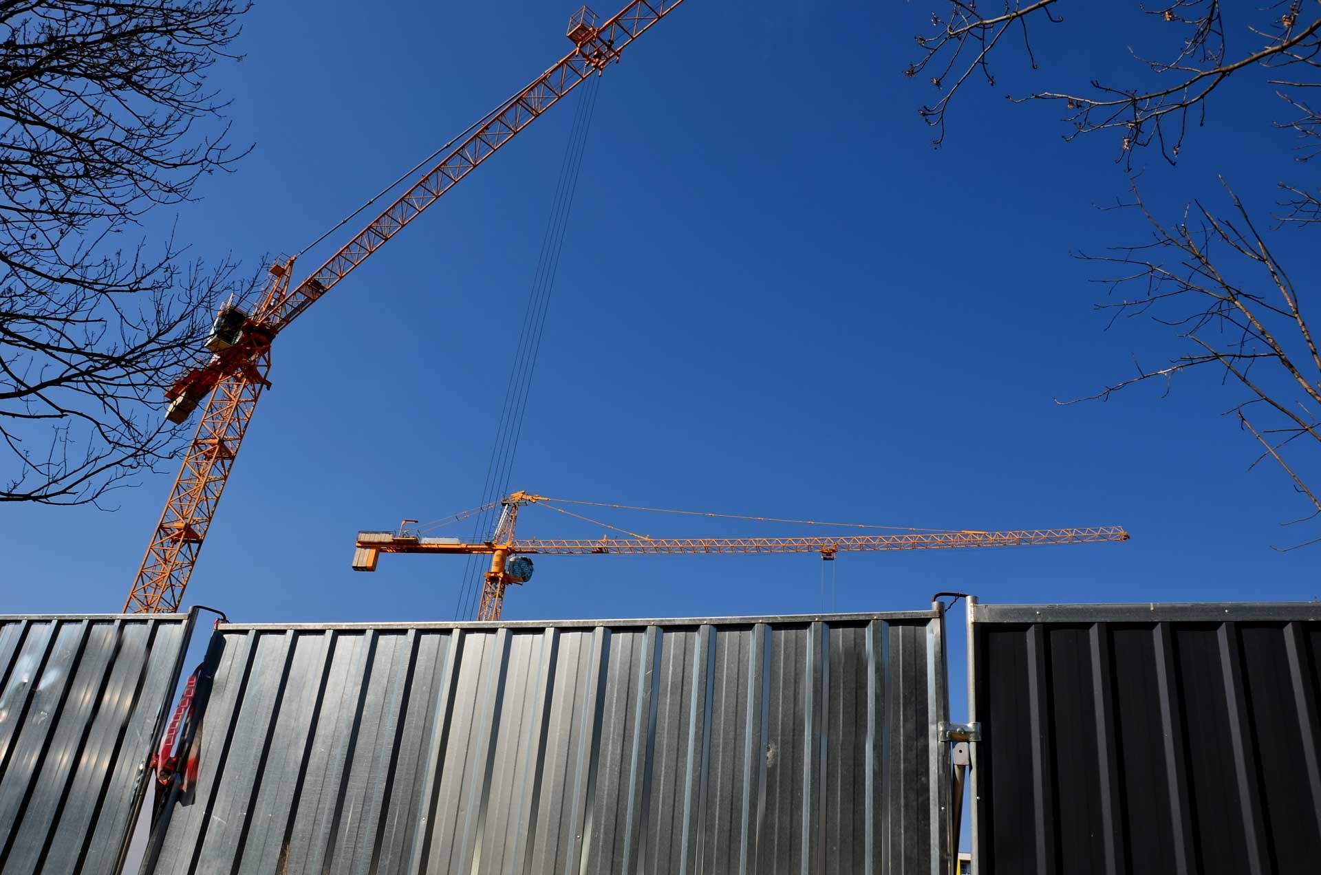 Two orange tower cranes stand behind a metal fence beneath a clear blue sky. Two orange tower cranes stand behind a metal fence beneath a clear blue sky.