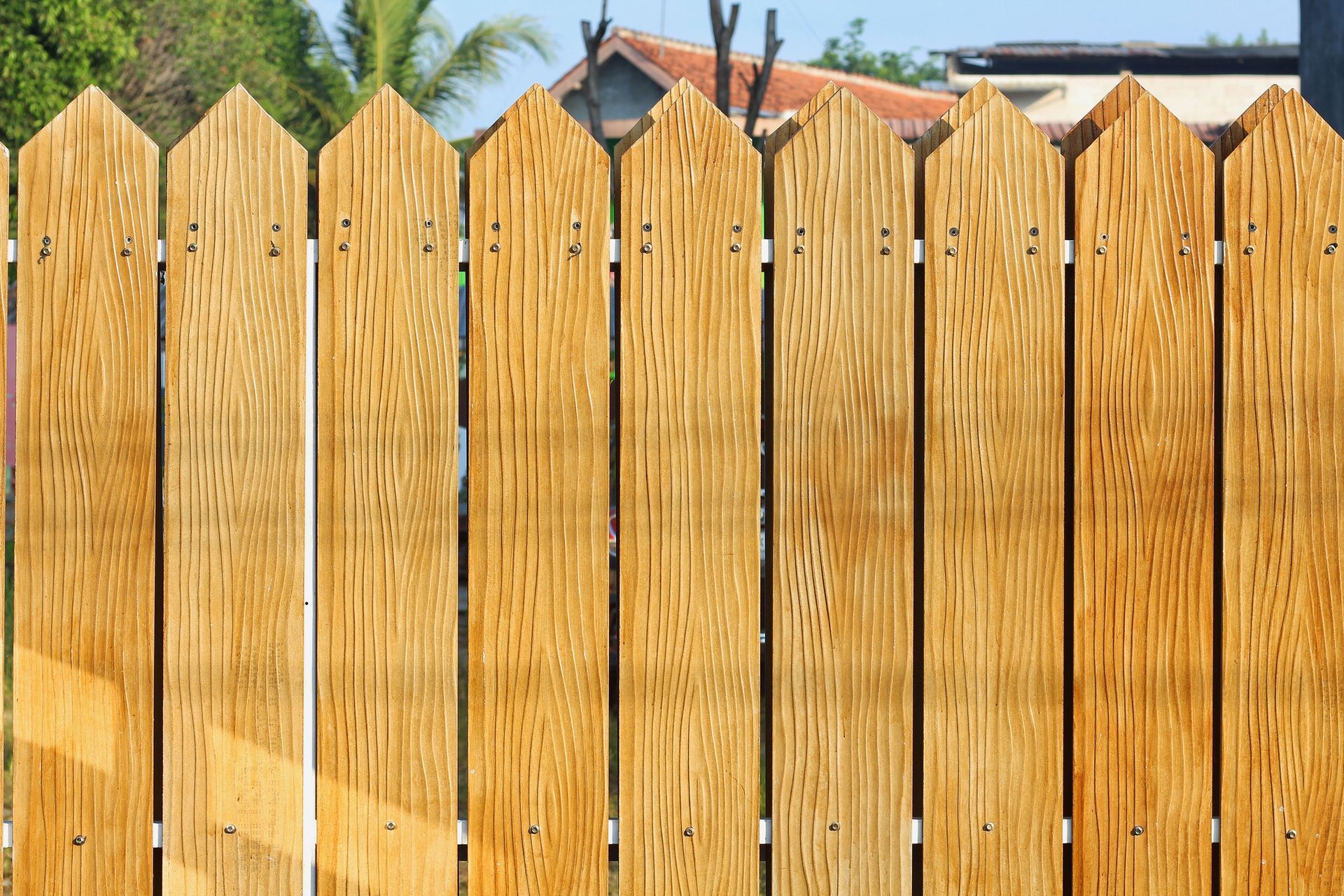 Wooden picket fence with pointed tops, set against trees and rooftops.