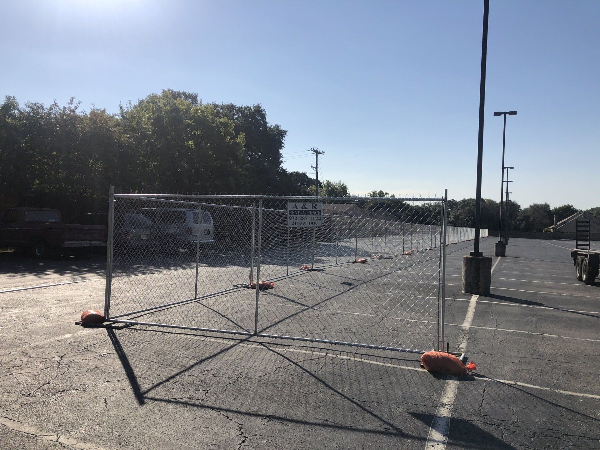 Chain-link fence in a parking lot, orange bases, bright sun, and trees in the background.