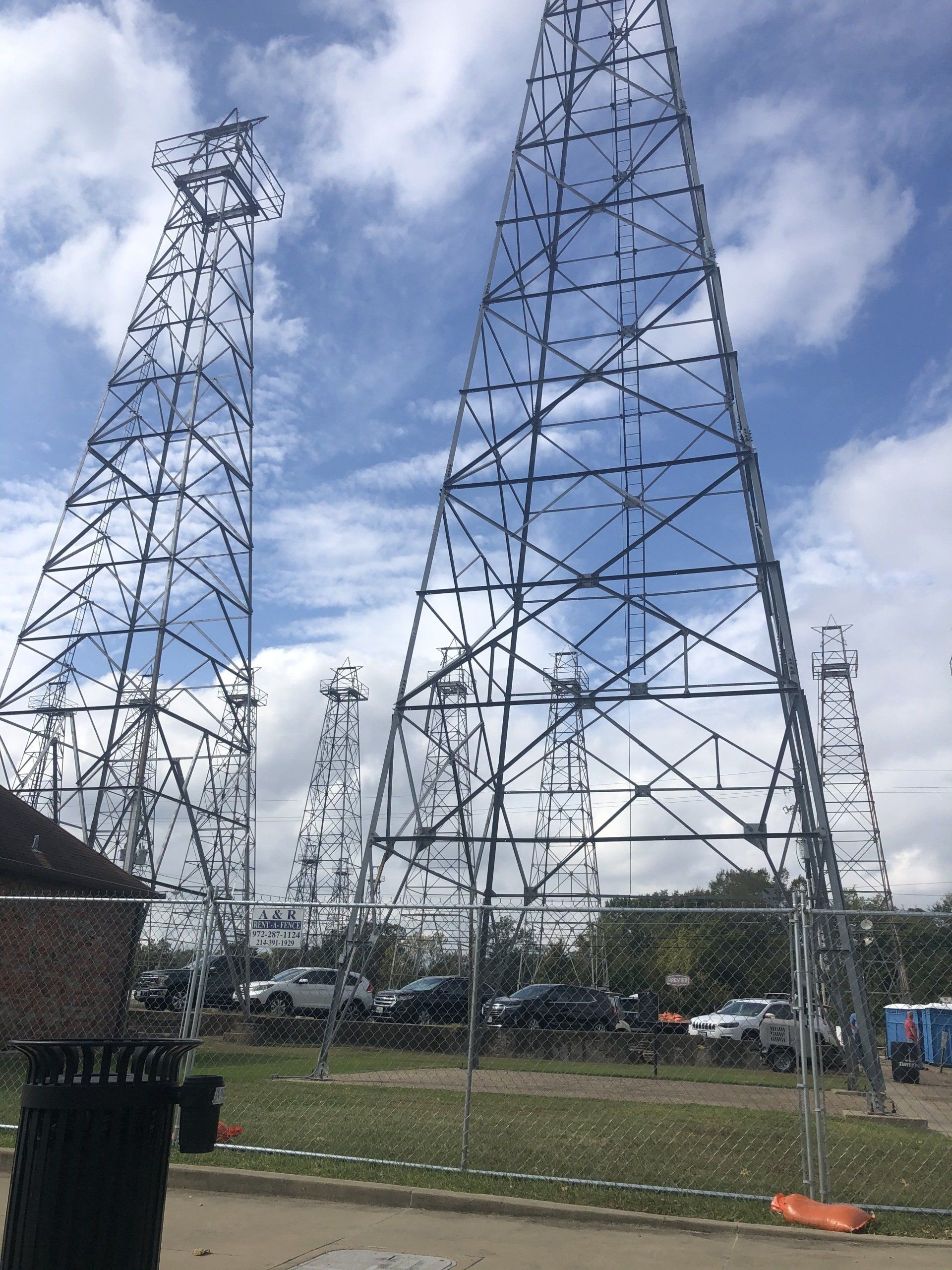 Tall, open-frame metal towers under a cloudy sky. Cars and greenery are visible at their base.