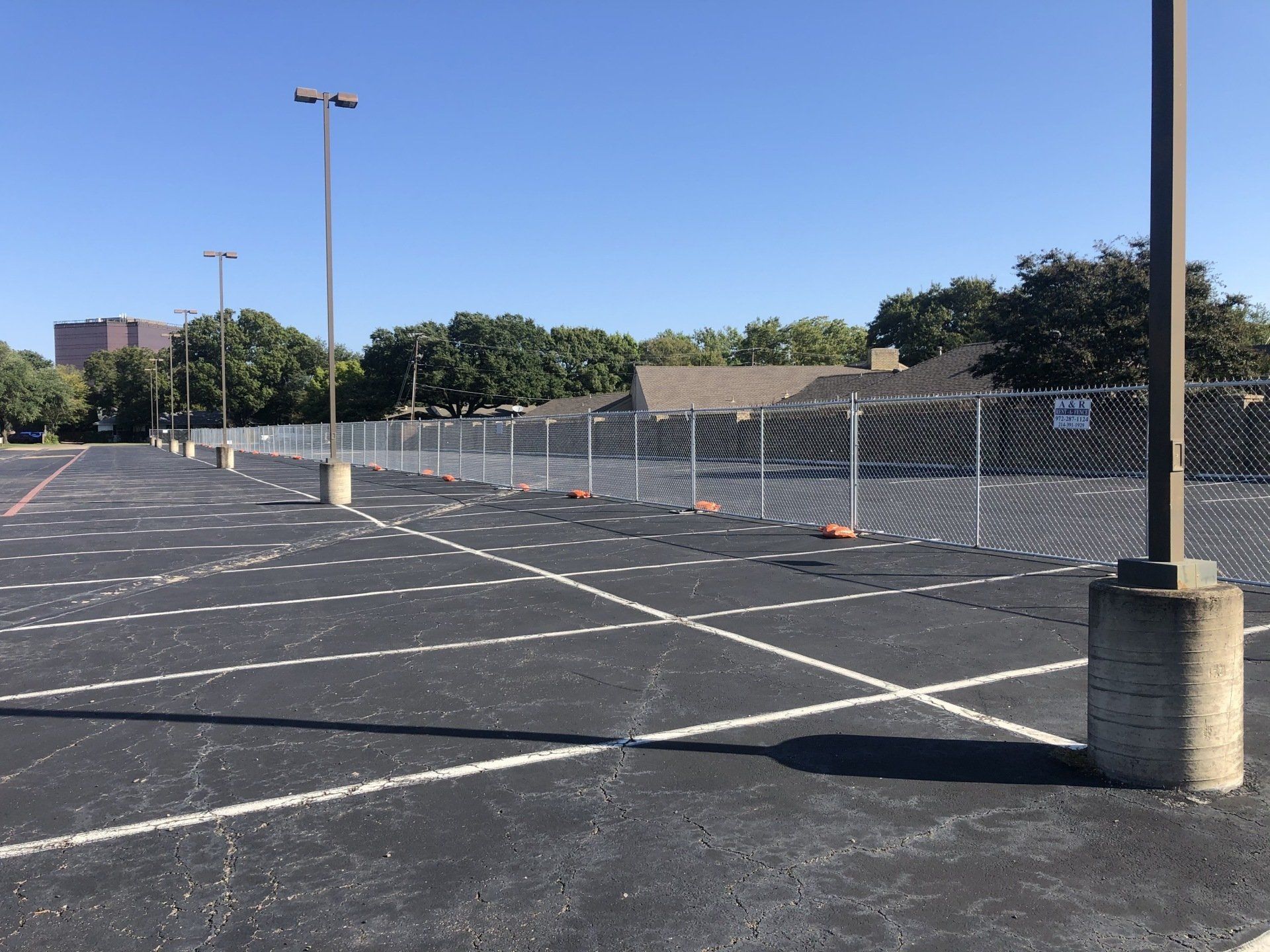 Empty parking lot with a chain-link fence in front of trees and a building on a sunny day.