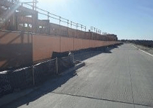 Construction site with long, rusty-colored barrier beside a concrete road. Clear, sunny sky.