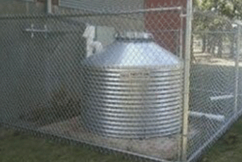 Metal water storage tank behind a chain-link fence on a concrete pad, surrounded by grass.