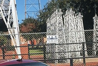 Model towers behind a chain-link fence; a red car is in view, and a tall structure is in the background.