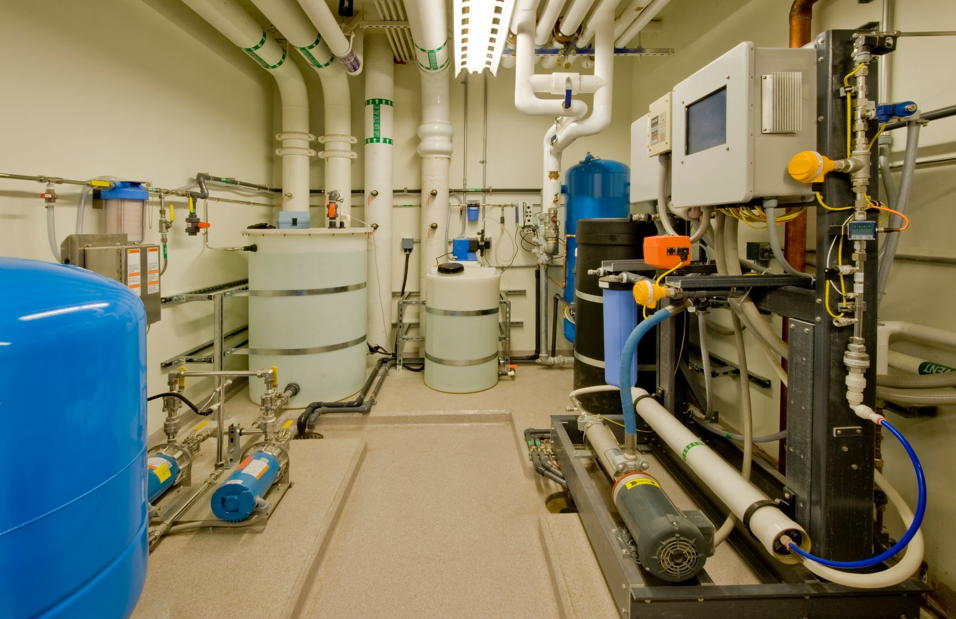 Water treatment room with pipes, tanks, and control panels. Blue and white equipment on a beige floor.