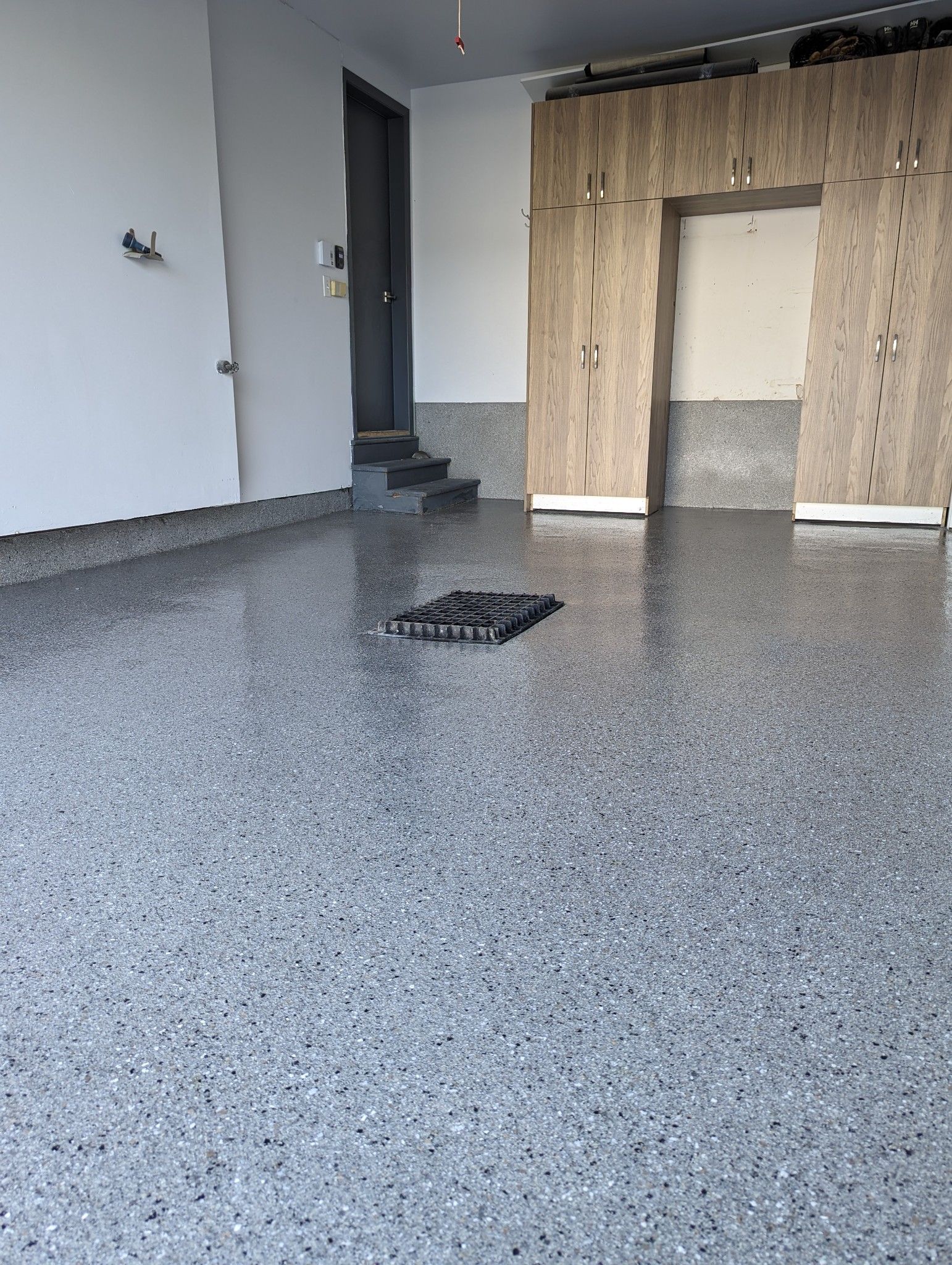 Plancher de garage en époxy gris moucheté avec une porte foncée et un mur partiellement recouvert.